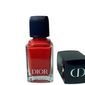 May include: A bottle of Dior nail polish in a vibrant red hue, with a black cap and the brand name printed on the front. The cap is off to the side, revealing the iconic CD logo.
