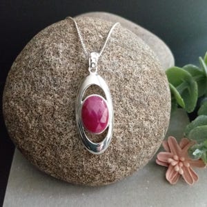 May include: A silver pendant necklace featuring a polished oval frame that holds a faceted, deep pinkish-red oval gemstone in the centre. The pendant hangs from a delicate silver chain, showcasing a smooth, reflective metallic finish.