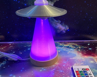 May include: A silver UFO shaped lamp with a purple light and a remote control with colour options. The lamp is on a table with a galaxy print.