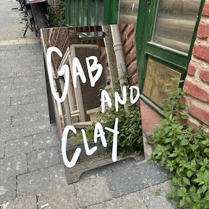 May include: A reflective sandwich board sign with the words "GAB AND CLAY" in white, handwritten-style lettering. The sign reflects the surrounding environment, including a brick wall, green door, and foliage. The sign is placed on a brick-paved sidewalk.