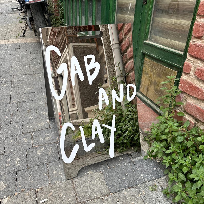 May include: A reflective sandwich board sign with the words "GAB AND CLAY" in white, handwritten-style lettering. The sign reflects the surrounding environment, including a brick wall, green door, and foliage. The sign is placed on a brick-paved sidewalk.
