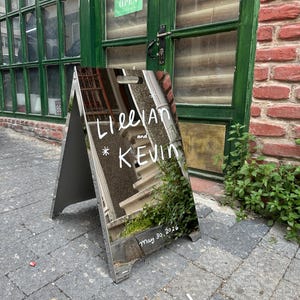 Peut inclure: Panneau en A avec surface réfléchissante. Le panneau affiche les noms "Lillian et Kevin" en écriture blanche, ainsi que la date "May 30, 2026." Le panneau est posé sur un trottoir pavé de briques, reflétant l'environnement.