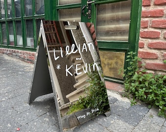 May include: A-frame sign with reflective surface. The sign displays the names "Lillian and Kevin" in white script, along with the date "May 30, 2026." The sign is positioned on a brick-paved walkway, reflecting the surrounding environment.
