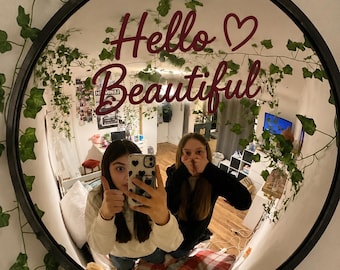 May include: A round black-framed wall mirror featuring the text Hello Beautiful in dark red script with a heart symbol. The mirror surface reflects a room interior with artificial green ivy vines draped around the outer edge of the circular frame.