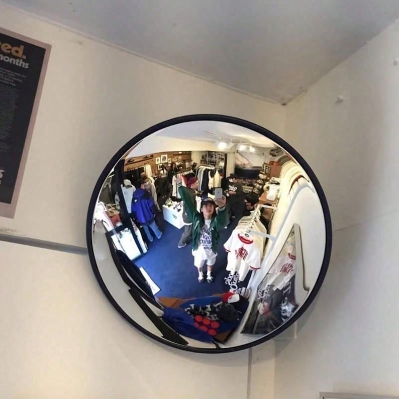 May include: A circular convex security mirror with a black frame mounted on a white wall. The mirror reflects a retail store interior with clothing racks, shelves, and individuals browsing merchandise, creating a wide-angle, distorted view.