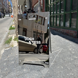 May include: A reflective wedding sign with the text "THE WEDDING OF JOSH & TESS" and the date "MAY 16 2026". The sign is displayed on a city street, reflecting the surroundings.
