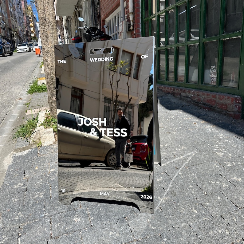 May include: A reflective wedding sign with the text "THE WEDDING OF JOSH & TESS" and the date "MAY 16 2026". The sign is displayed on a city street, reflecting the surroundings.