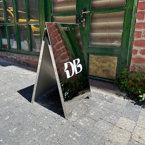May include: A silver A-frame sign with the white letters "DB" on the front. The sign is reflective, mirroring the surroundings. It stands on a brick-paved surface near a green door and brick wall.
