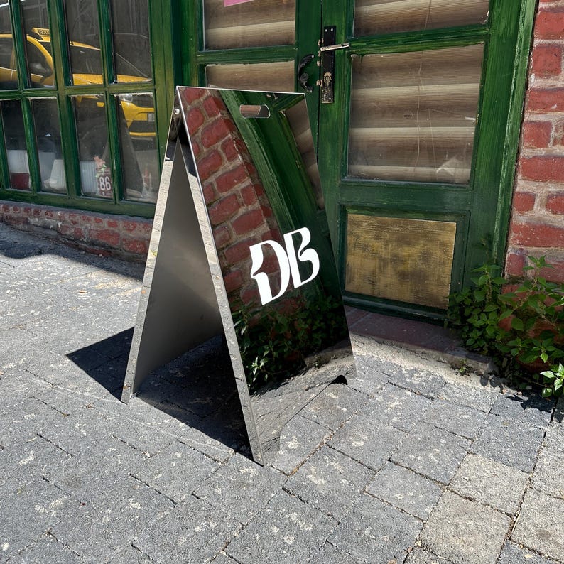 May include: A silver A-frame sign with the white letters "DB" on the front. The sign is reflective, mirroring the surroundings. It stands on a brick-paved surface near a green door and brick wall.