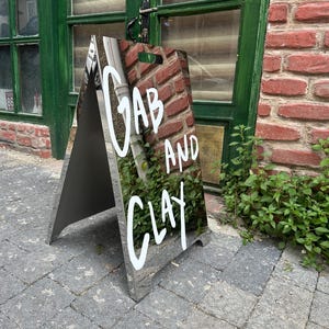 May include: A-frame sign with the words "GAB AND CLAY" in white lettering. The sign has a reflective surface and is positioned on a brick-paved sidewalk in front of a green door and brick wall. The sign is angled towards the viewer.
