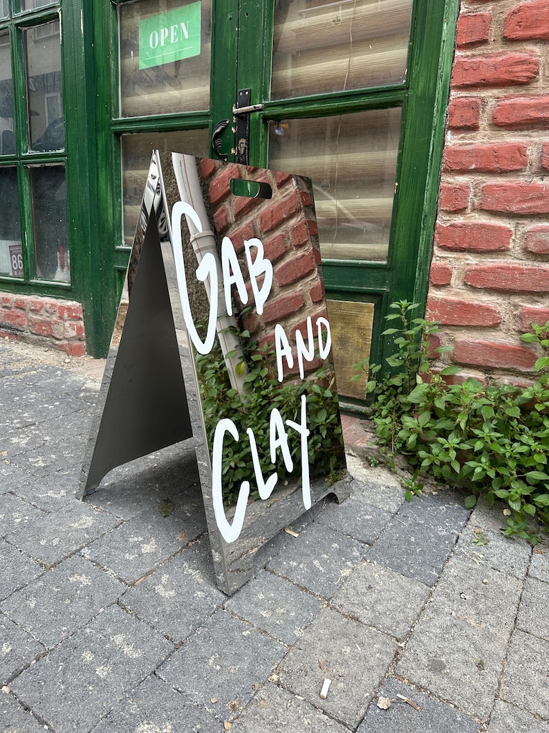 May include: A-frame sign with the words "GAB AND CLAY" in white lettering. The sign has a reflective surface and is positioned on a brick-paved sidewalk in front of a green door and brick wall. The sign is angled towards the viewer.