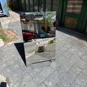 May include: A mirrored wedding sign with the text "THE WEDDING OF JOSH & TESS" and the date "MAY 16, 2026". The sign reflects the surrounding street scene, including cars and buildings.