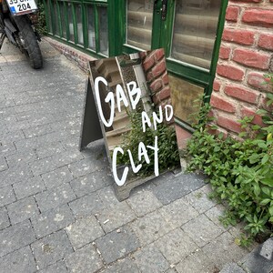 May include: An A-frame sign with the words "Gab and Clay" in white script. The sign is placed on a brick and stone surface, with a green door and foliage in the background.