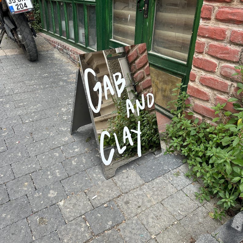 May include: An A-frame sign with the words "Gab and Clay" in white script. The sign is placed on a brick and stone surface, with a green door and foliage in the background.