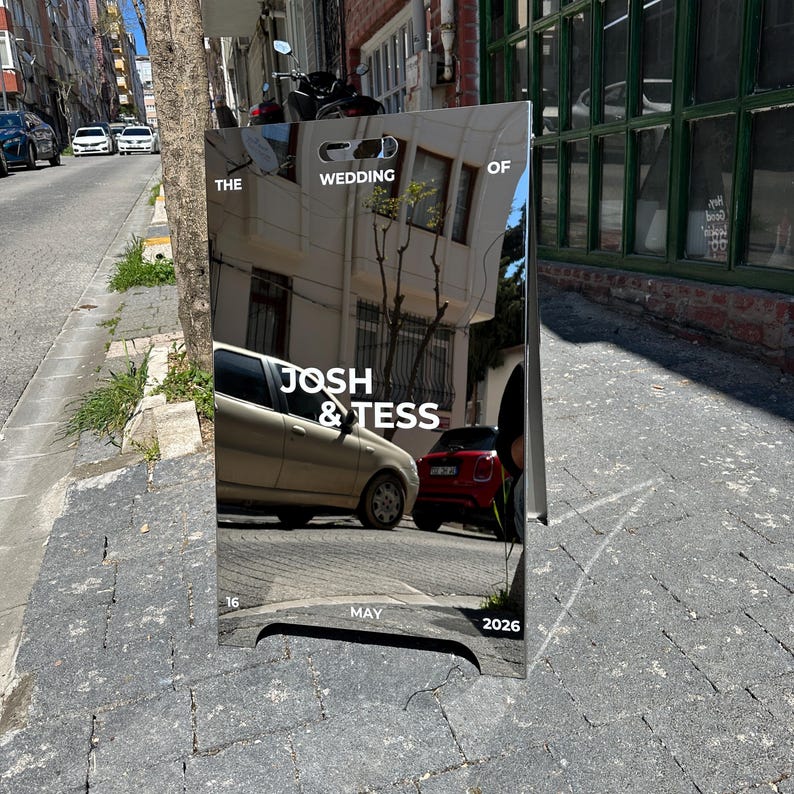 May include: A reflective wedding sign with the text "THE WEDDING OF JOSH & TESS" and the date "MAY 16 2026". The sign reflects the street scene, including cars and buildings.