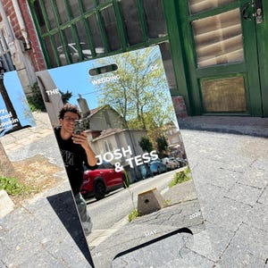 May include: A rectangular mirror with a handle at the top, reflecting a street scene and a person taking a selfie. The text "THE WEDDING OF JOSH & TESS" is printed on the mirror, along with "MAY 2026".