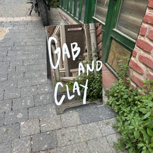 May include: A-frame sign with the words "Gab and Clay" in white script. The sign is reflective, showing a blurred image of a building and greenery. The sign is on a brick-paved sidewalk, with a motorcycle tire visible in the background.