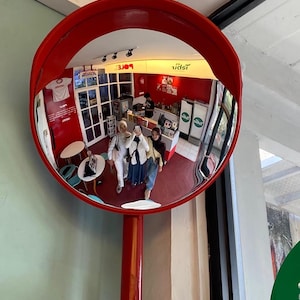 May include: A large, circular, convex security mirror with a bright red frame and a matching red support pole. The mirror reflects an interior space with red walls, white tables, and three individuals standing together while taking a photograph.