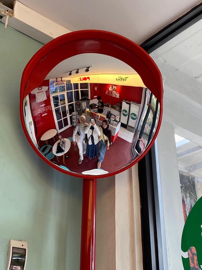 May include: A large, circular, convex security mirror with a bright red frame and a matching red support pole. The mirror reflects an interior space with red walls, white tables, and three individuals standing together while taking a photograph.