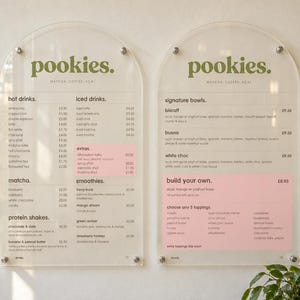 May include: Two arched clear acrylic menu boards with olive green text titled pookies. The left board lists hot drinks, iced drinks, matcha, and protein shakes. The right board lists signature bowls and build your own options with pink accents.