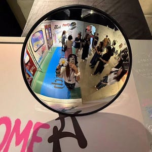 May include: A circular convex security mirror with a black frame reflecting a room filled with people, graffiti-covered walls, and a blue floor. The reflection shows a person holding a white flower-shaped accessory while taking a photo.