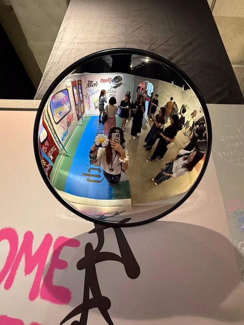 May include: A circular convex security mirror with a black frame reflecting a room filled with people, graffiti-covered walls, and a blue floor. The reflection shows a person holding a white flower-shaped accessory while taking a photo.