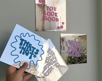 May include: Four square mirror tiles featuring colorful vinyl lettering and flower accents. One mirror displays blue text reading THATS HOT, another pink text YOU LOOK GOOD, a third purple text I JUST DONT CARE, and the fourth text MANIFEST AND WORK.