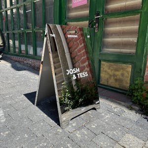 Pode incluir: Um letreiro de casamento em forma de A com o texto "THE WEDDING OF JOSH & TESS MAY 16 2026". O letreiro apresenta uma fotografia de uma parede de tijolos, escadas e vegetação. O letreiro é cinzento e está sobre uma superfície de tijolo.