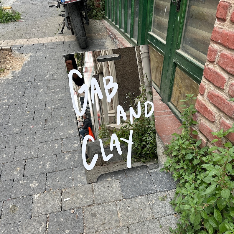 May include: A reflective sign with the words "GAB AND CLAY" in white script. The sign is propped up on a brick-paved sidewalk, with a motorcycle and building visible in the background. The sign reflects the surrounding environment.