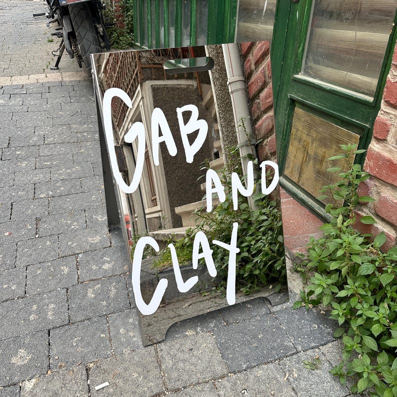 May include: A reflective sign with the words "GAB AND CLAY" in white script. The sign is propped up on a brick and cobblestone surface, with a glimpse of a building reflected in the mirror.