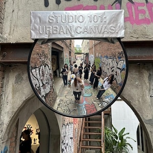 May include: A circular convex mirror mounted on a brick wall with graffiti, featuring a silver rectangular sign above that reads STUDIO 101: URBAN CANVAS. The mirror reflects an alleyway with people and colorful street art on the surrounding walls.