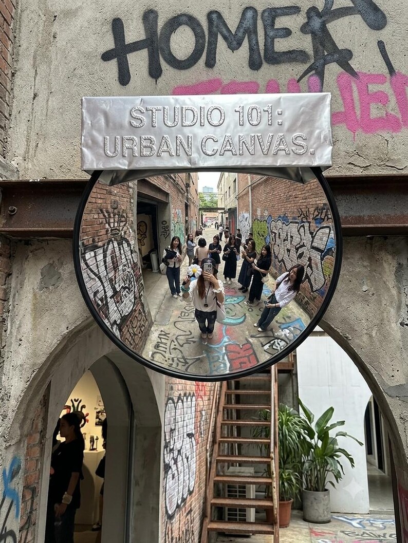 May include: A circular convex mirror mounted on a brick wall with graffiti, featuring a silver rectangular sign above that reads STUDIO 101: URBAN CANVAS. The mirror reflects an alleyway with people and colorful street art on the surrounding walls.