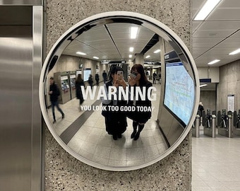 May include: A circular convex security mirror with a white frame mounted on a concrete wall. The mirror surface displays the white text WARNING YOU LOOK TOO GOOD TODAY and a smaller plaque below reading STATION SECURITY / SELF-OBSERVATION POINT.
