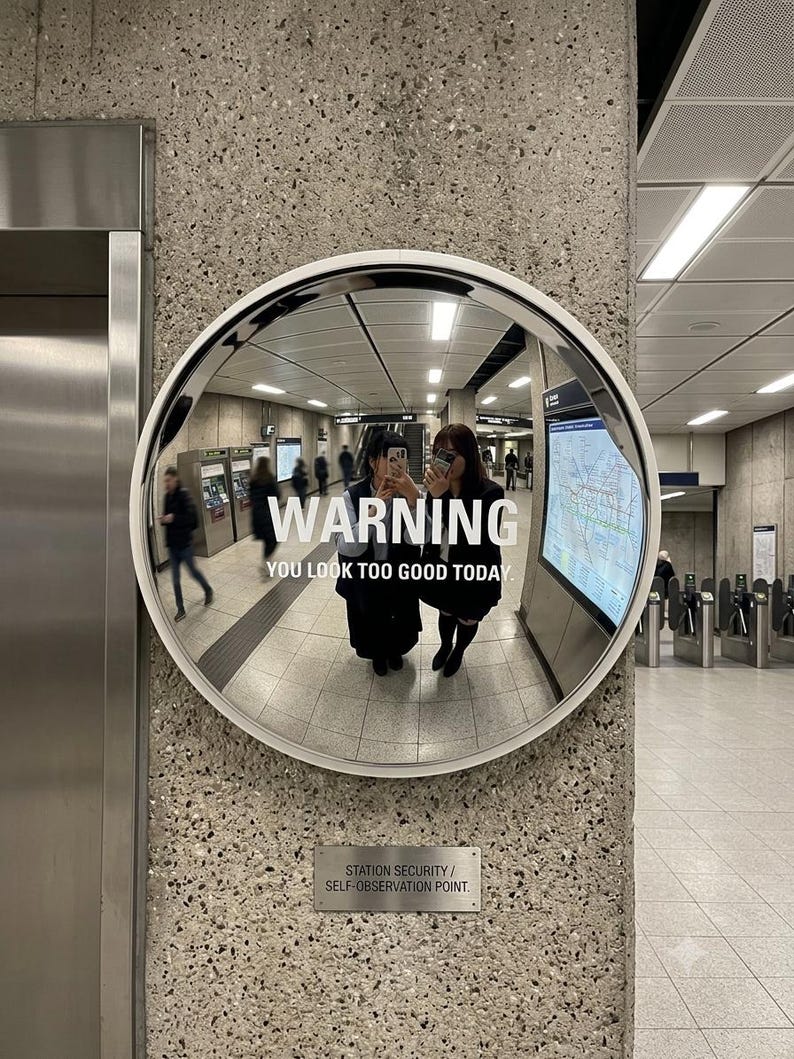 May include: A circular convex security mirror with a white frame mounted on a concrete wall. The mirror surface displays the white text WARNING YOU LOOK TOO GOOD TODAY and a smaller plaque below reading STATION SECURITY / SELF-OBSERVATION POINT.