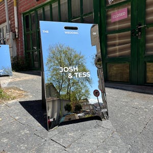 May include: A mirrored wedding sign with the text "THE WEDDING OF JOSH & TESS MAY 16 2026". The sign reflects a tree, buildings, and a blue sky. The sign is an A-frame style.