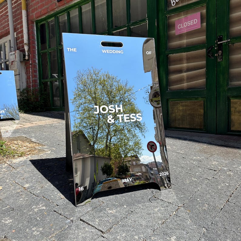 May include: A mirrored wedding sign with the text "THE WEDDING OF JOSH & TESS MAY 16 2026". The sign reflects a tree, buildings, and a blue sky. The sign is an A-frame style.