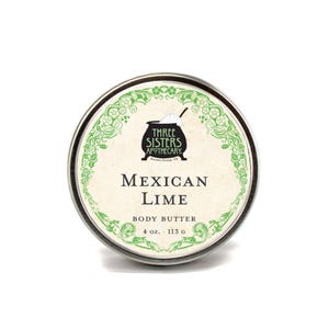 May include: A silver tin with a green and white label. The label features a black cauldron with the text "Three Sisters Apothecary" and the words "Mexican Lime Body Butter 4 oz. 113 g".
