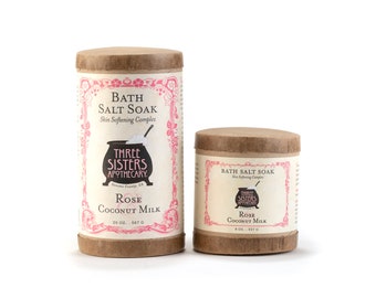 Op de afbeelding: Twee bruine kartonnen containers met badzout met roze en zwarte etiketten. De grotere container is 20 oz en de kleinere 8 oz. Beide containers zijn gelabeld met "Bath Salt Soak" en "Three Sisters Apothecary". De grotere container is gelabeld met "Rose Coconut Milk" en de kleinere met "Rose Coconut Milk".