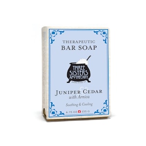 May include: A blue and white bar soap package with a black and white illustration of a cauldron. The text on the package reads "Therapeutic Bar Soap", "Three Sisters Apothecary", "Sonoma County, CA", "Juniper Cedar with Arnica", "Soothing & Cooling", "4.75 OZ 135 G".