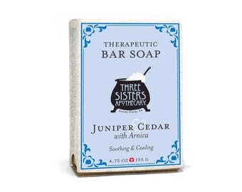 Op de afbeelding: Een blauwe en witte zeepverpakking met een zwart-witte illustratie van een ketel. De tekst op de verpakking luidt "Therapeutic Bar Soap", "Three Sisters Apothecary", "Sonoma County, CA", "Juniper Cedar with Arnica", "Soothing & Cooling", "4.75 OZ 135 G".