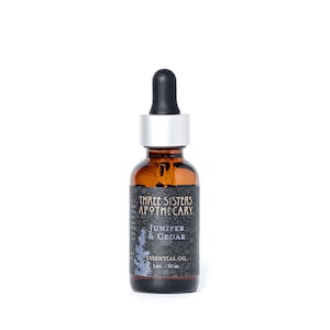 May include: A brown glass bottle with a silver cap and dropper. The bottle contains Juniper & Cedar essential oil from Three Sisters Apothecary. The label is black with white text and a small illustration of a pine tree. The bottle contains 30 ml of oil.