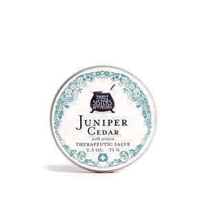 May include: A round, silver tin of Juniper Cedar Therapeutic Salve by Three Sisters Apothecary. The label features a teal floral design and the text "Juniper Cedar with arnica Therapeutic Salve 2.5 oz." The tin also includes a medical cross symbol.