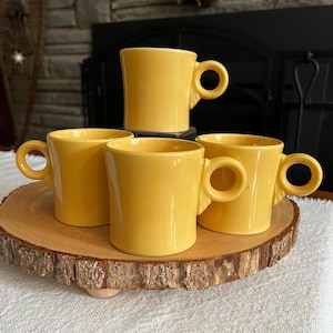 Puede incluir: Cuatro tazas de cerámica amarillas con asas redondeadas dispuestas sobre una bandeja de madera. Las tazas son de un amarillo brillante y tienen un diseño sencillo. La bandeja tiene un borde natural y rústico.