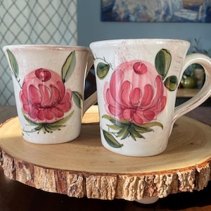 May include: Two ceramic mugs with a floral design. The mugs are white with a pink flower and green leaves painted on them. They are sitting on a round wooden serving tray.
