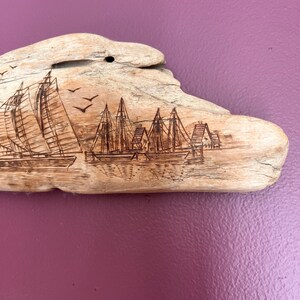 May include: Driftwood art piece featuring etched sailboats and buildings. The natural wood has a light brown color, with the etched details appearing darker. Three birds are etched in flight above the sailboats. The piece is mounted on a purple background.
