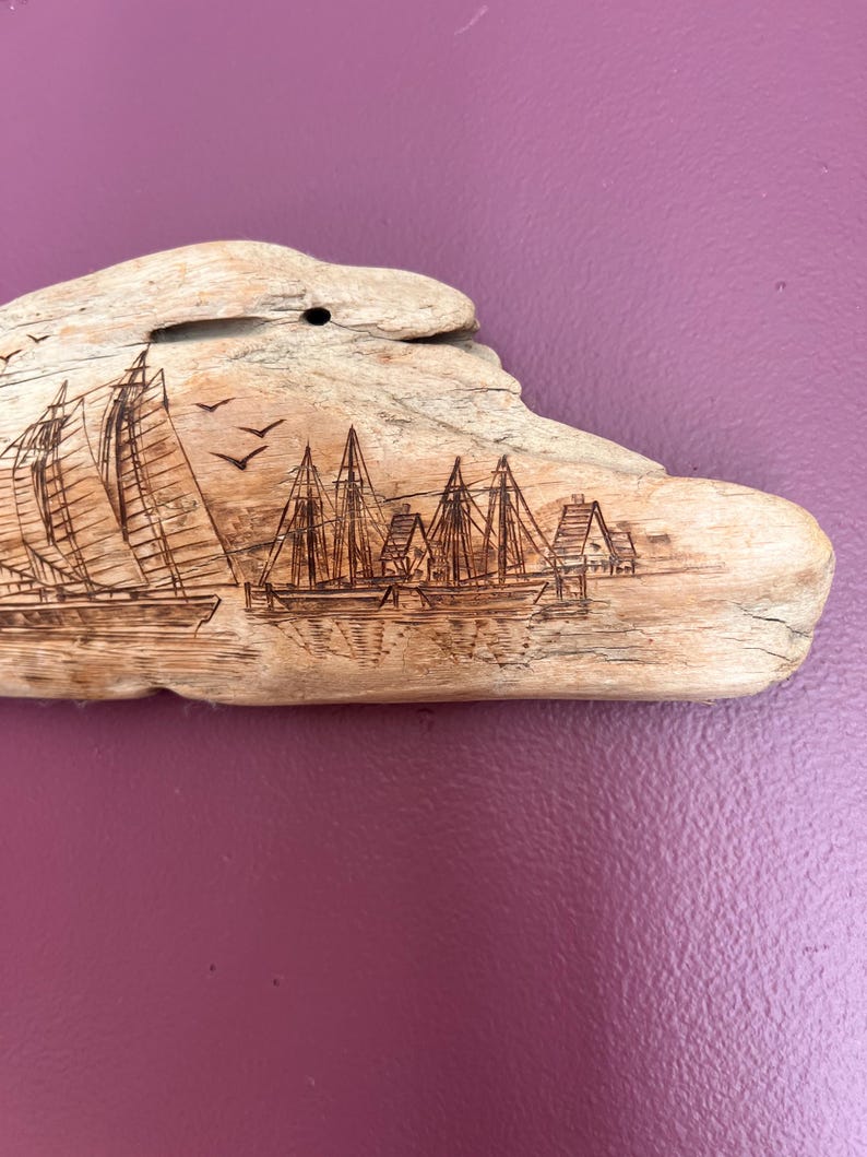 May include: Driftwood art piece featuring etched sailboats and buildings. The natural wood has a light brown color, with the etched details appearing darker. Three birds are etched in flight above the sailboats. The piece is mounted on a purple background.