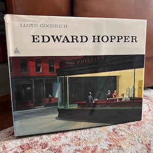 May include: A book cover featuring the painting "Nighthawks" by Edward Hopper. The cover has the title "EDWARD HOPPER" in large black letters, with the artist's name, Lloyd Goodrich, above. The painting depicts a diner scene.