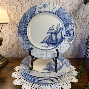 May include: A stack of blue and white nautical-themed plates featuring a map design and a sailboat illustration. The plates are displayed on a black metal stand and a white lace doily, creating a vintage aesthetic.