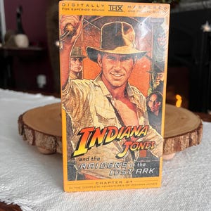 May include: A vintage VHS tape of "Indiana Jones and the Raiders of the Lost Ark." The cover features a portrait of the main character in a fedora and tan shirt. The title is in a bold, stylized font, with the THX logo at the top.