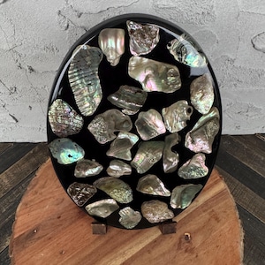 May include: A black, oval-shaped decorative piece inlaid with iridescent abalone shells. The shells vary in size and shape, displaying a range of colors including silver, green, and pink. The piece is displayed on a wooden stand.